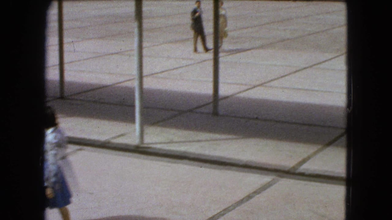 Pedestrians cross a sunlit tiled plaza behind vertical posts.