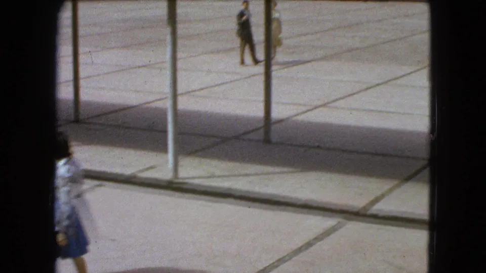 In Mexico City, 1962, pedestrians move across a bright tiled plaza with vertical posts and long shadows in the foreground.