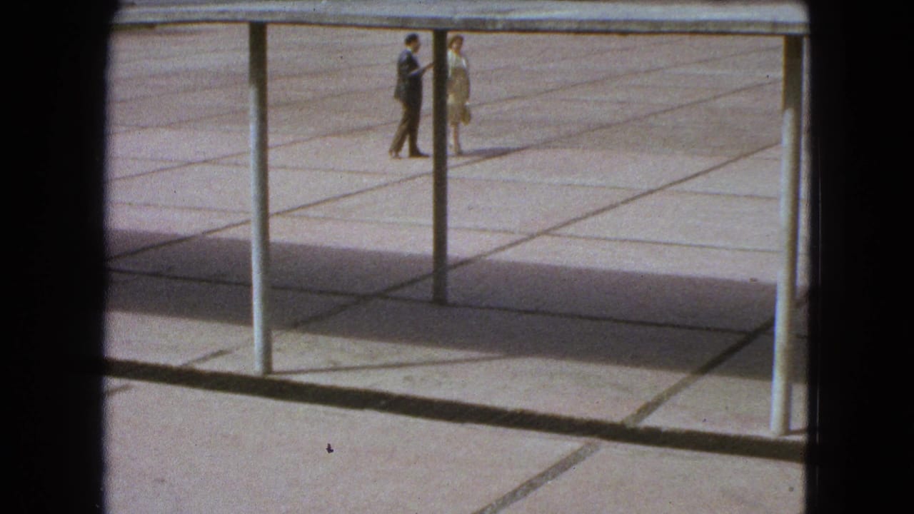 Two pedestrians walk across a paved plaza beside vertical posts.
