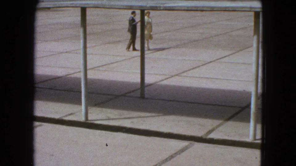 Two distant pedestrians walk through a sunlit Mexico City plaza in 1962, with tiled paving and structural posts visible.
