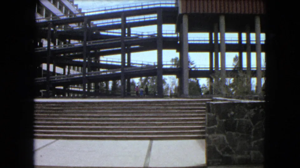 A modern Mexico City architectural complex appears with elevated walkways, concrete supports, broad stairs, and plaza paving.