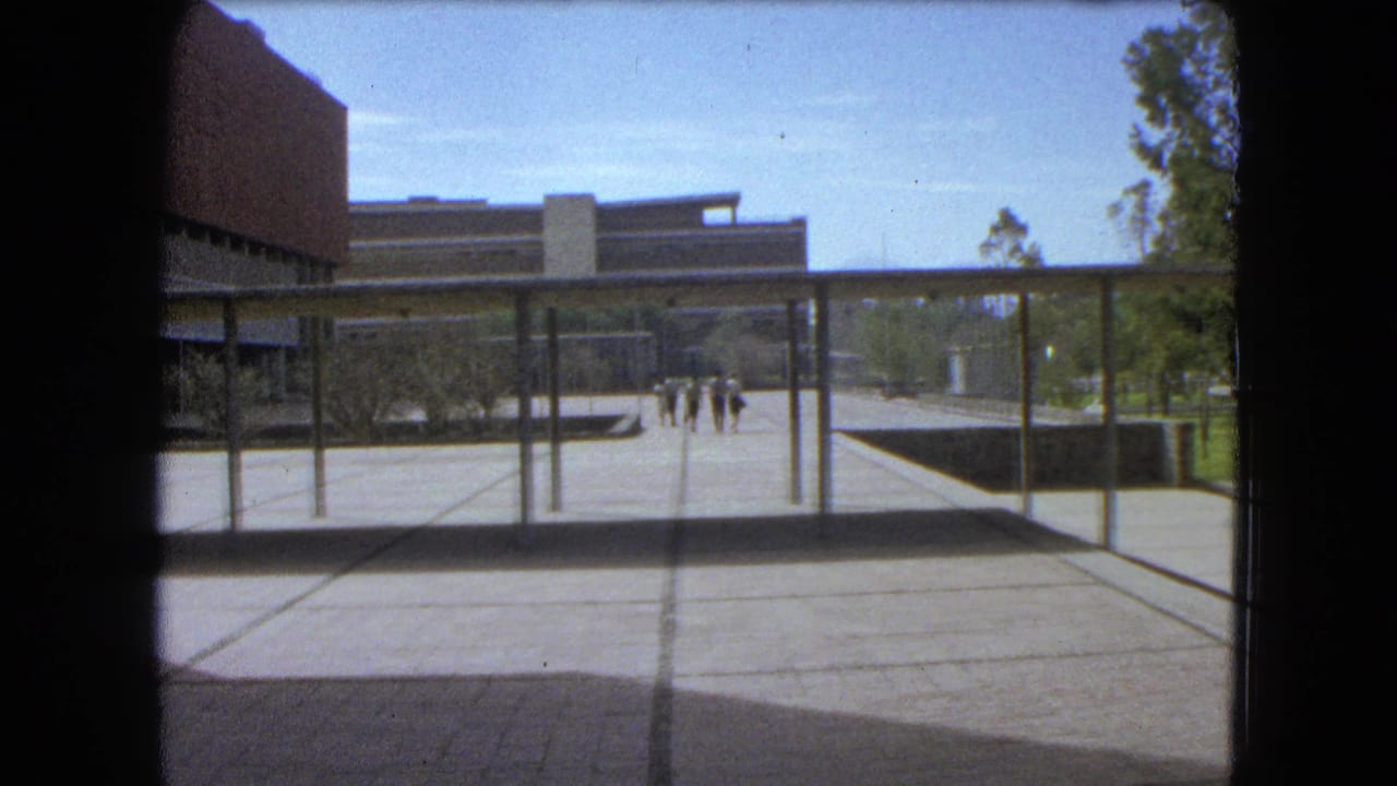 1962 Mexico City 8mm frame showing a modern courtyard, covered walkway, distant pedestrians, trees, and dark scan borders.