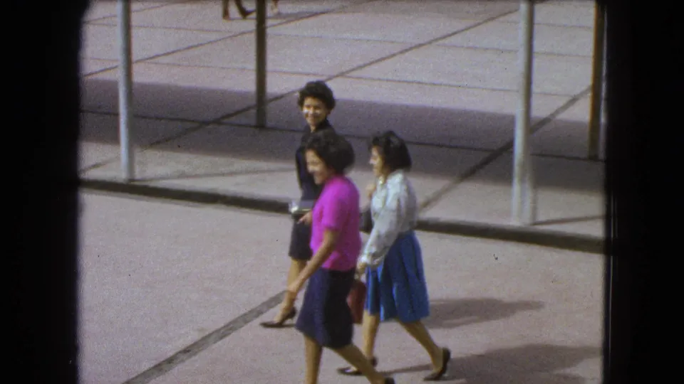 In 1962 Mexico City footage, three women continue across the courtyard as one glances toward the camera.
