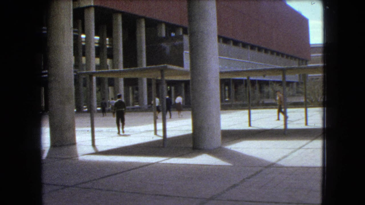 1962 Mexico City home-movie frame showing people crossing a paved courtyard near a brick building with tall concrete columns and canopy structures.