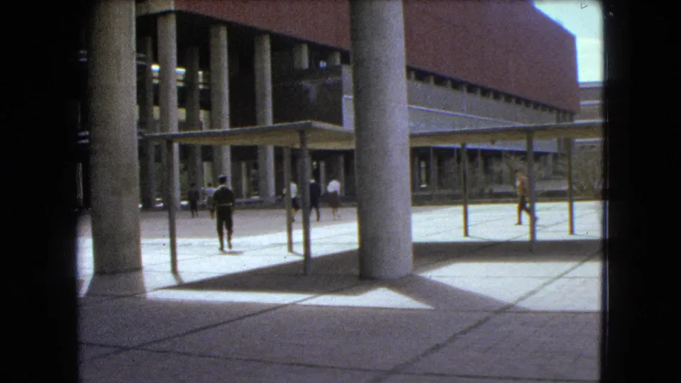 1962 Mexico City home-movie frame showing people crossing a paved courtyard near a brick building with tall concrete columns and canopy structures.