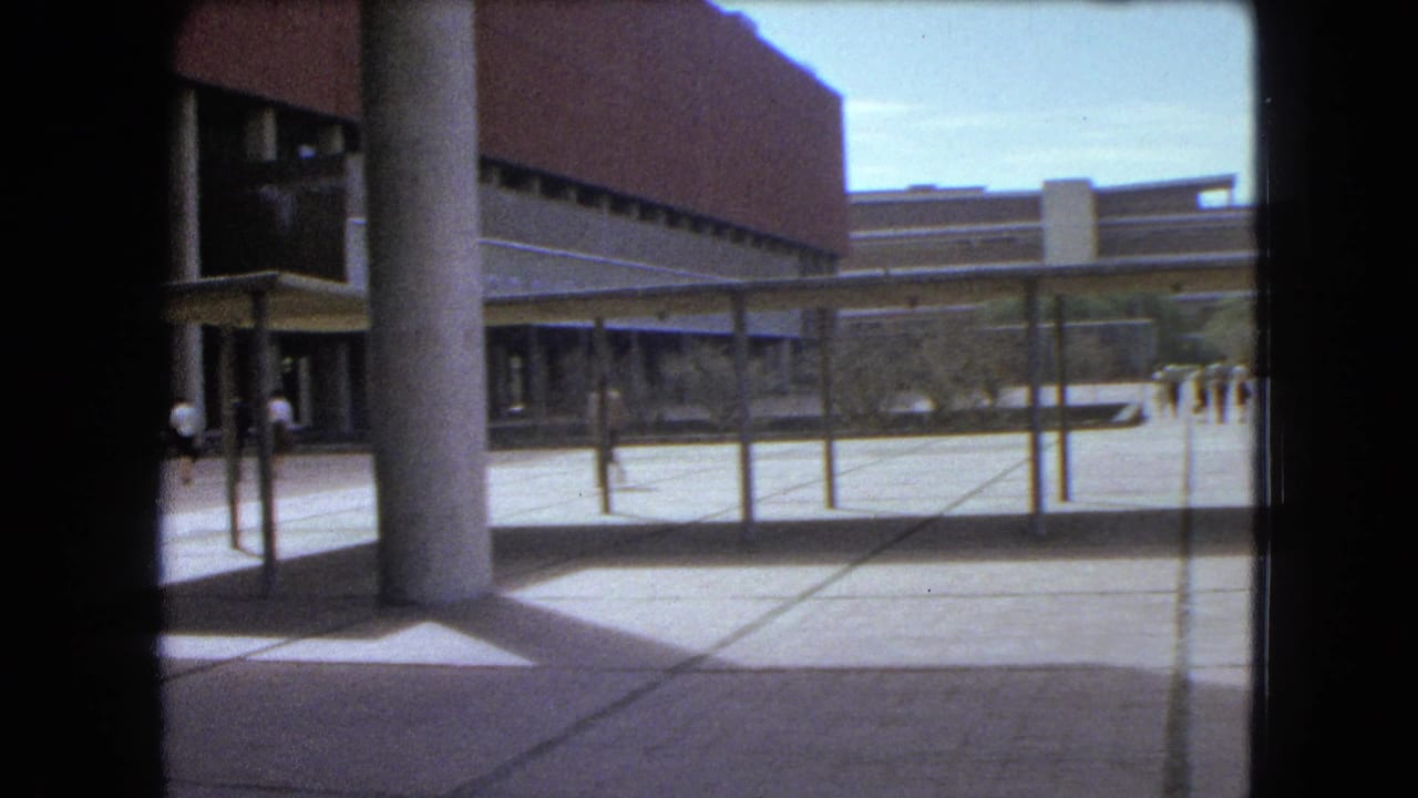 A paved plaza and covered walkway sit in front of a brick building with pedestrians in the distance.