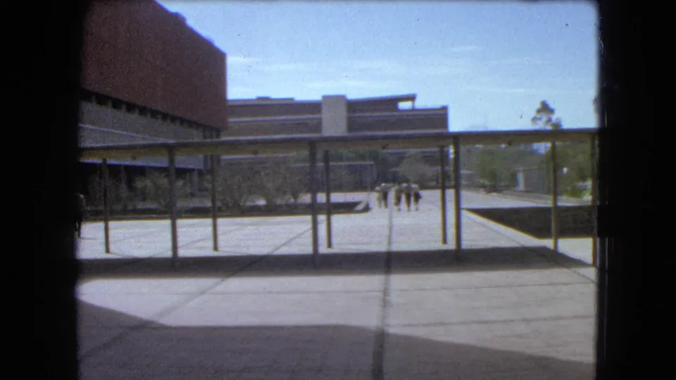 1962 Mexico City home-movie frame looking across a modernist plaza with canopy posts, brick architecture, trees, and small groups of pedestrians.