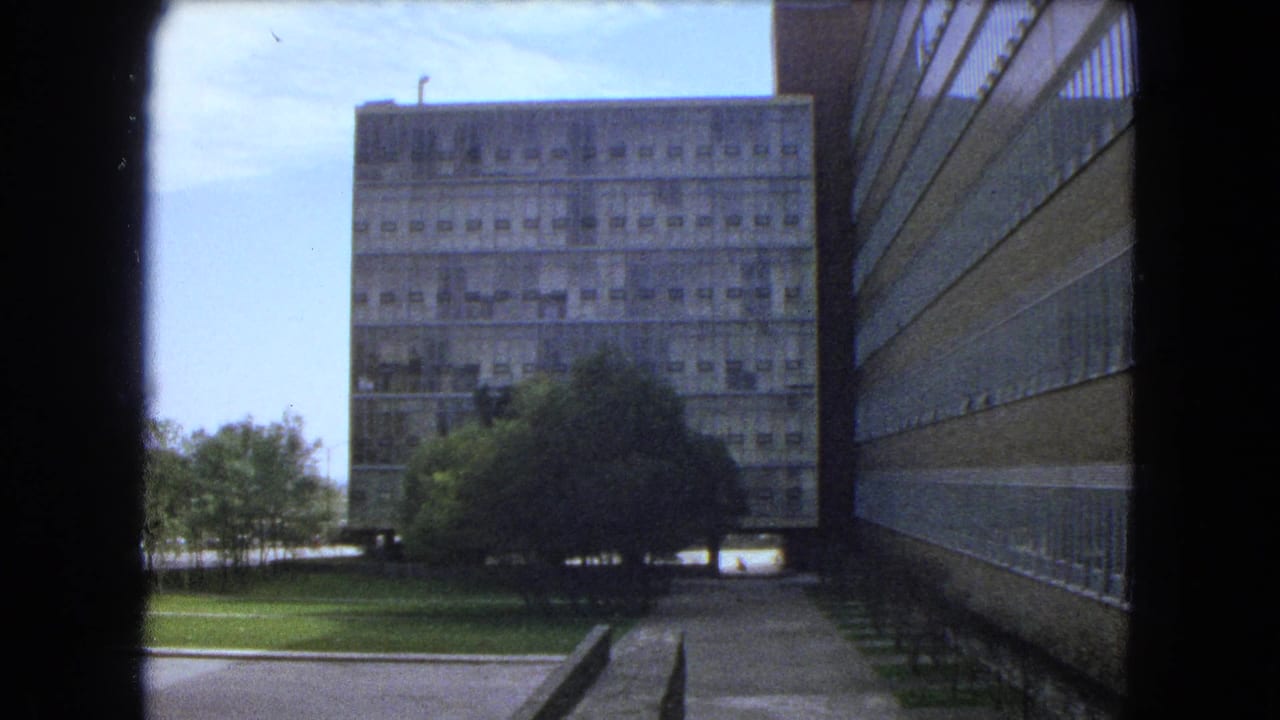 1962 Mexico City 8mm view of a large concrete building, lawn, trees, and a walkway seen from a shaded opening.