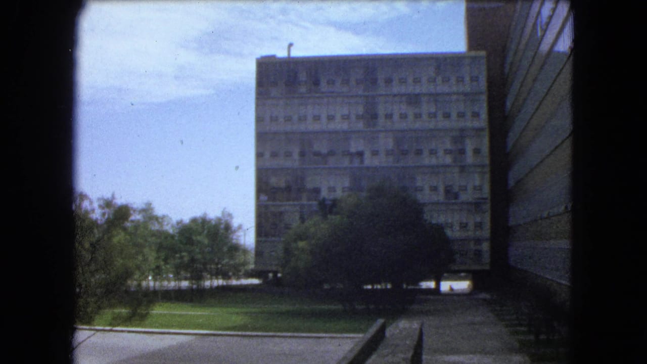 Concrete building and trees viewed past a dark vertical frame and side wall.