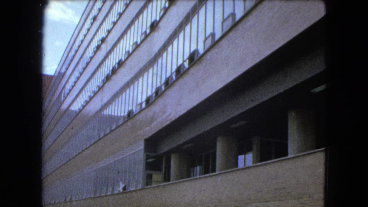 Low-angle view along a concrete and glass high-rise with a second building partly visible behind it.