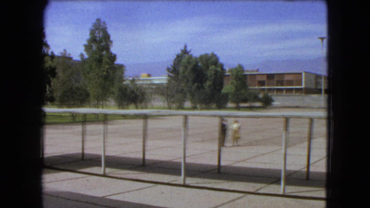 1962 Mexico City school campus view with a paved courtyard, railings, trees, and distant figures near a modern building.