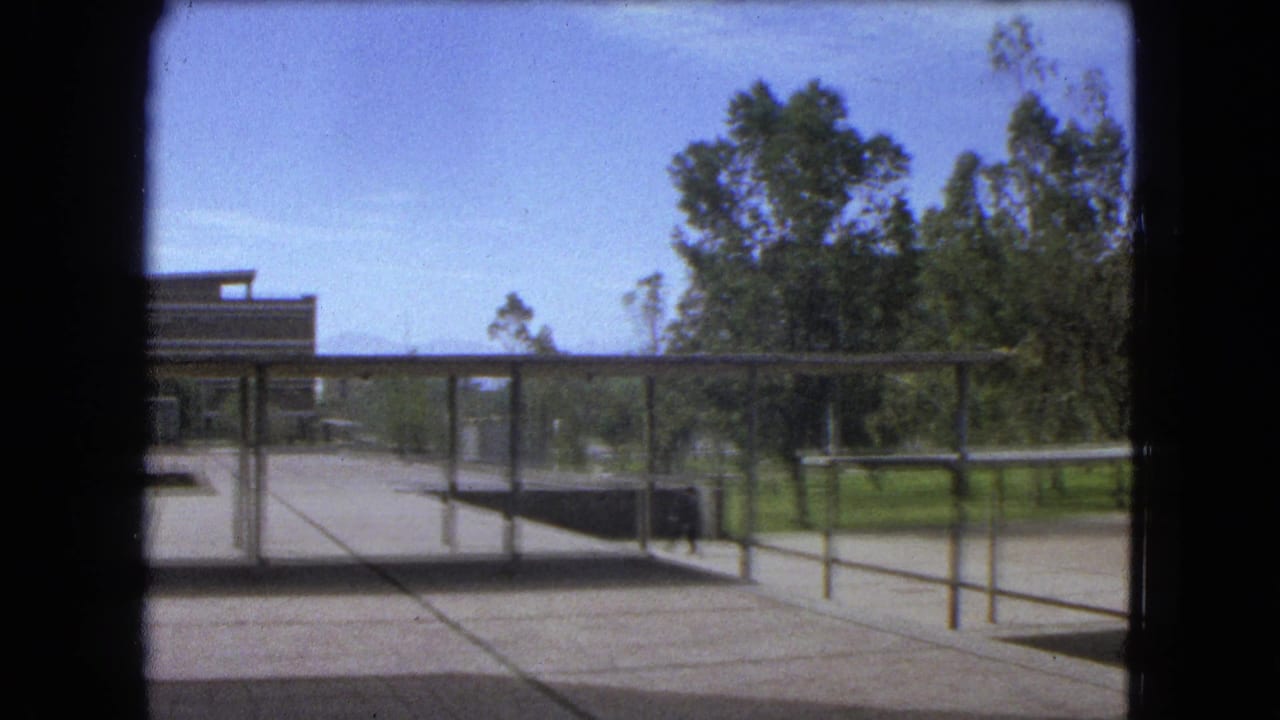 Concrete campus plaza with metal fencing, open shelter, trees, and a partial building at left.