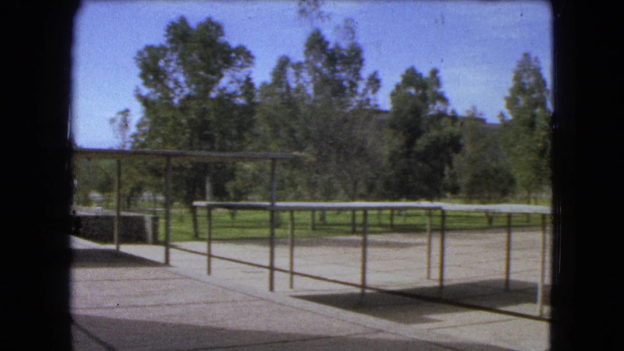 Open paved area with metal railings, shade structure, trees, lawn, and bright sky.