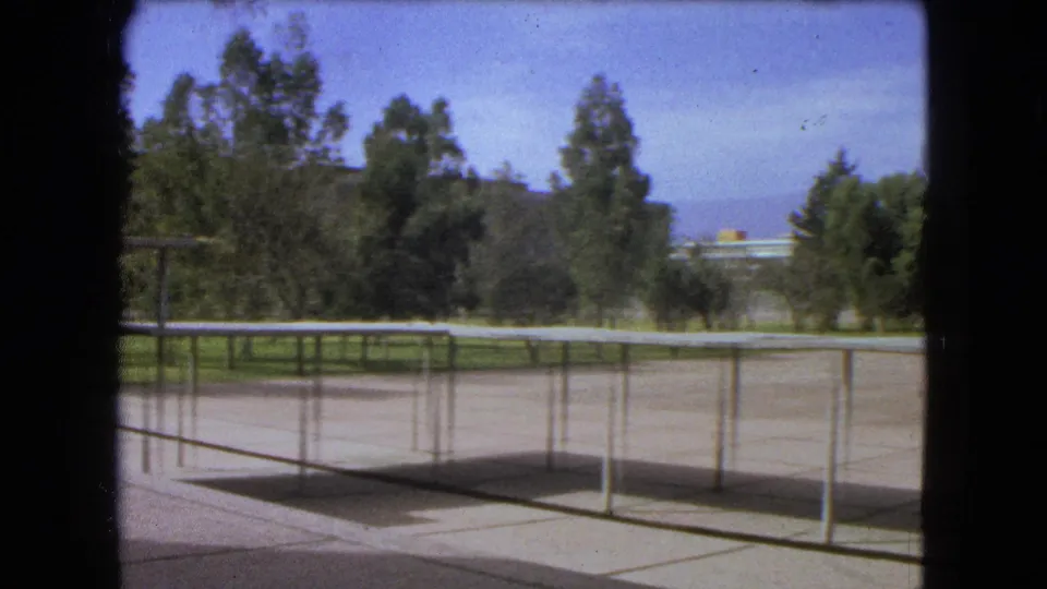 1962 Mexico City 8mm frame with a fence-lined campus plaza, trees, grass, a distant building, and hazy hills in the background.