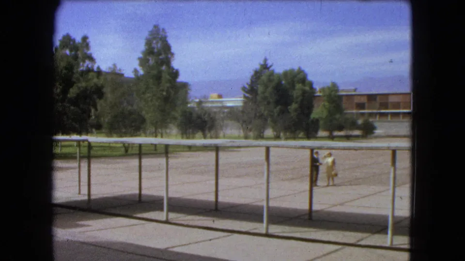 1962 Mexico City 8mm frame showing an open campus plaza with railings, trees, a distant building, and two people walking at right.