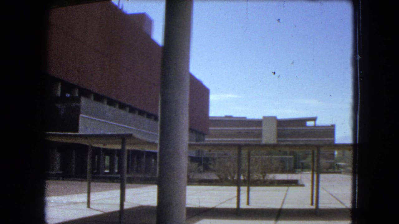 1962 Mexico City courtyard view through a dark frame, showing brick and concrete architecture, a column, paved ground, and clear sky.
