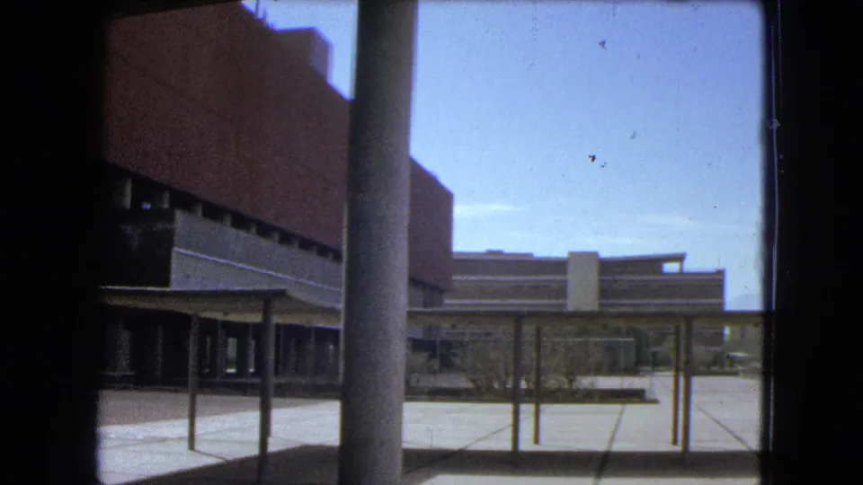 1962 Mexico City courtyard view through a dark frame, showing brick and concrete architecture, a column, paved ground, and clear sky.