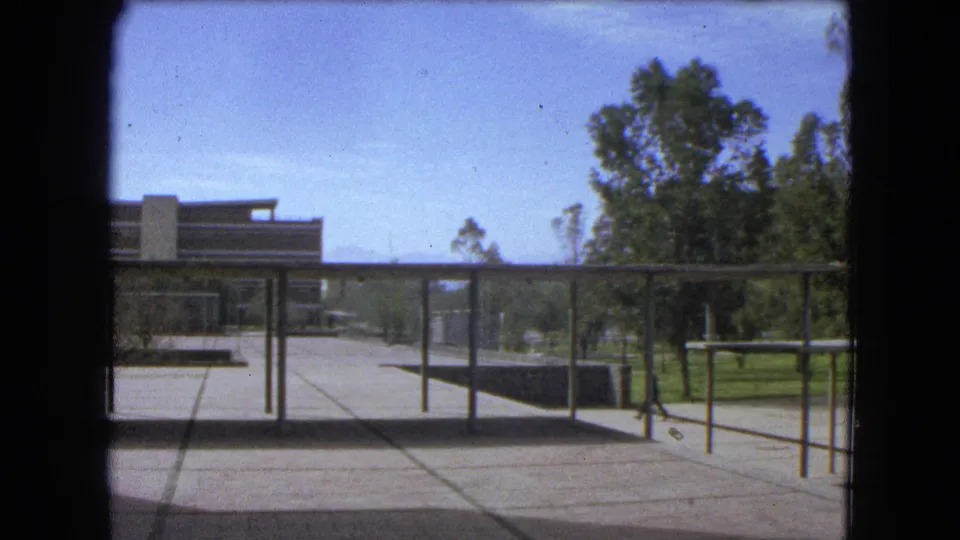 1962 Mexico City courtyard with modern building forms, walkway posts, trees, lawn, and a small kite-like object against the sky.