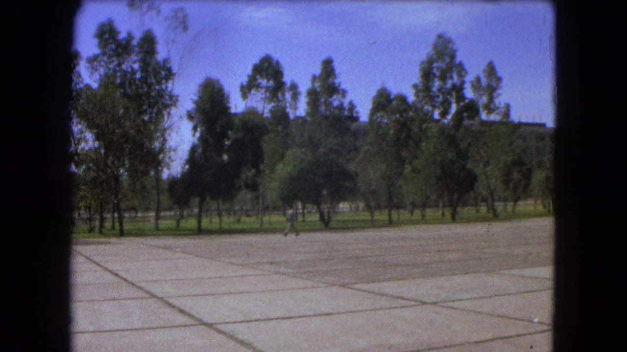 1962 Mexico City home-movie frame showing a distant student crossing a broad concrete plaza lined with trees.
