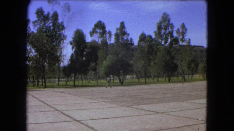 1962 Mexico City home-movie frame showing a distant student crossing a broad concrete plaza lined with trees.