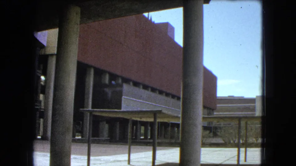 Columned exterior of a modern school building, seen from a shaded or window-framed home-movie viewpoint.
