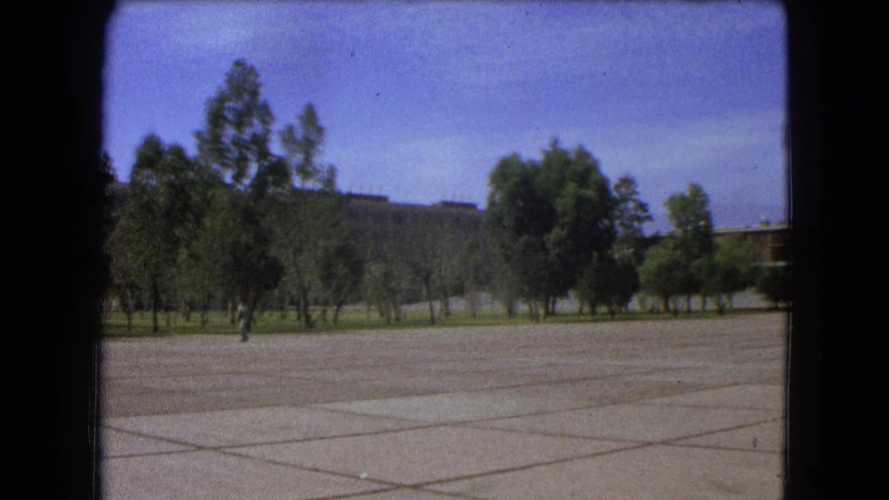 1962 Mexico City home-movie frame showing a broad empty paved plaza with trees, sky, and a distant man walking across the open space.