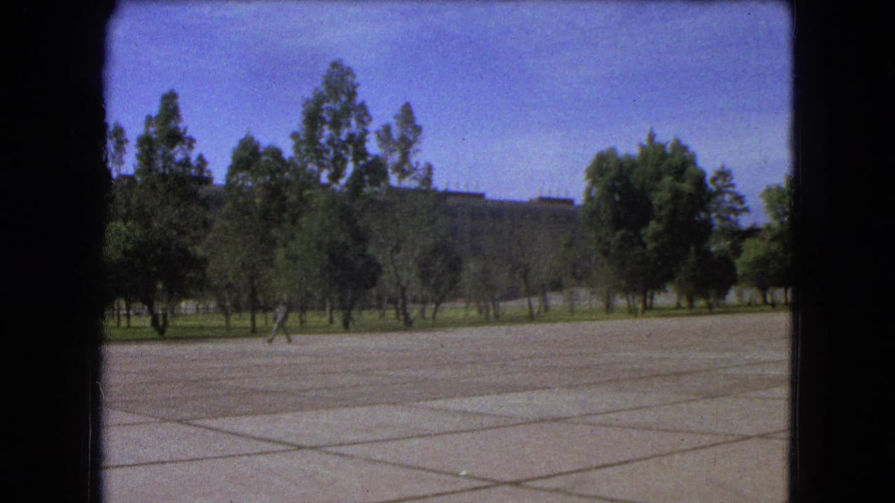 Man walking across an open paved plaza with trees and distant structures behind him.
