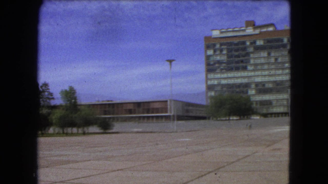 1962 Mexico City 8mm frame showing a broad paved plaza, modernist buildings, trees, a lamppost, and a lone distant walker.