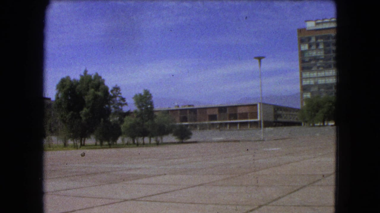 1962 Mexico City 8mm frame with an empty paved foreground, trees, low modernist building, lamppost, and partial high-rise.
