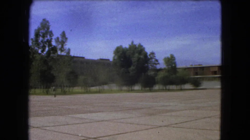1962 Mexico City 8mm frame showing an open paved plaza bordered by trees and modern buildings under a blue sky.