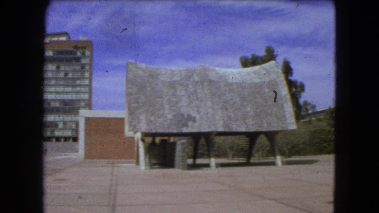 Wide plaza with a tall glass-and-brick building and a low textured-roof structure under a blue sky.