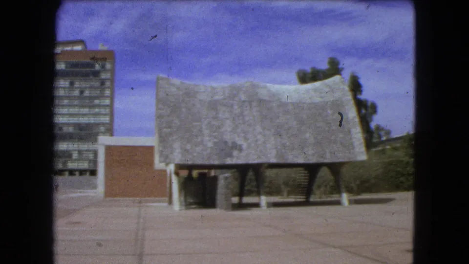 1962 Mexico City 8mm frame showing a paved plaza, modernist tower, and low sculptural-roof building from ground level.
