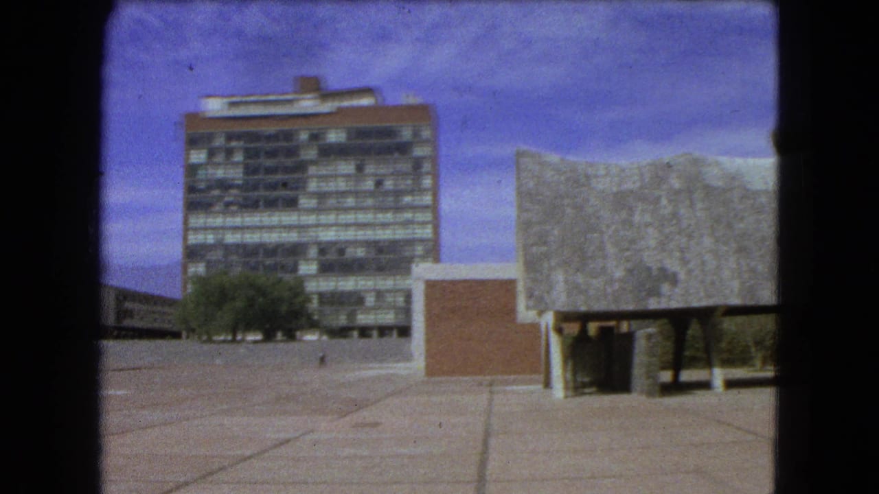 Modernist tower and low concrete-roofed building seen across an open paved plaza.