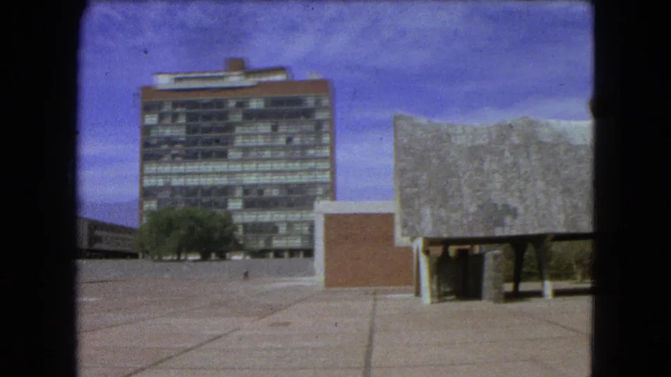 Mexico City exterior with a tall office-like block, brown wall section, trees, and a low textured-roof structure on pillars.