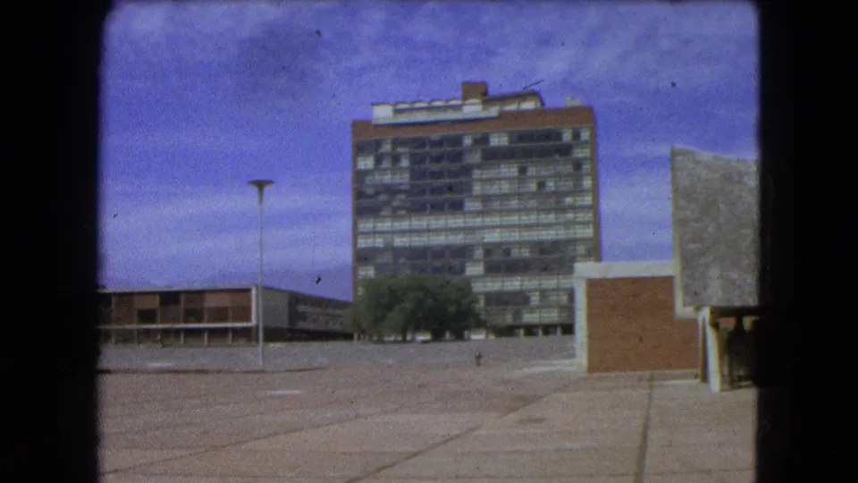 1962 Mexico City plaza view with a lamppost, glass-and-brick tower, low adjacent building, trees, and partial sculptural roof.