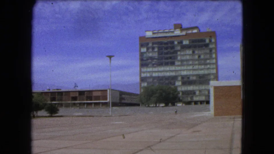 Final 8mm frame of a Mexico City modernist complex with open paving, lamppost, tower, trees, and blue sky.