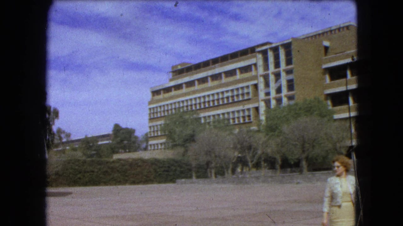 1962 Mexico City 8mm frame showing a modernist building, paved plaza, trees, hedges, and an adult woman crossing the foreground.