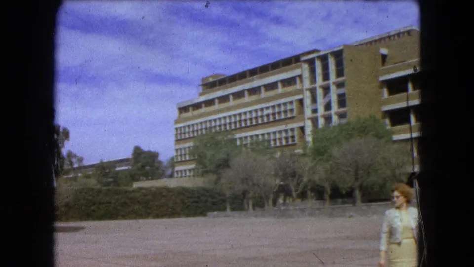 1962 Mexico City 8mm frame showing a modernist building, paved plaza, trees, hedges, and an adult woman crossing the foreground.
