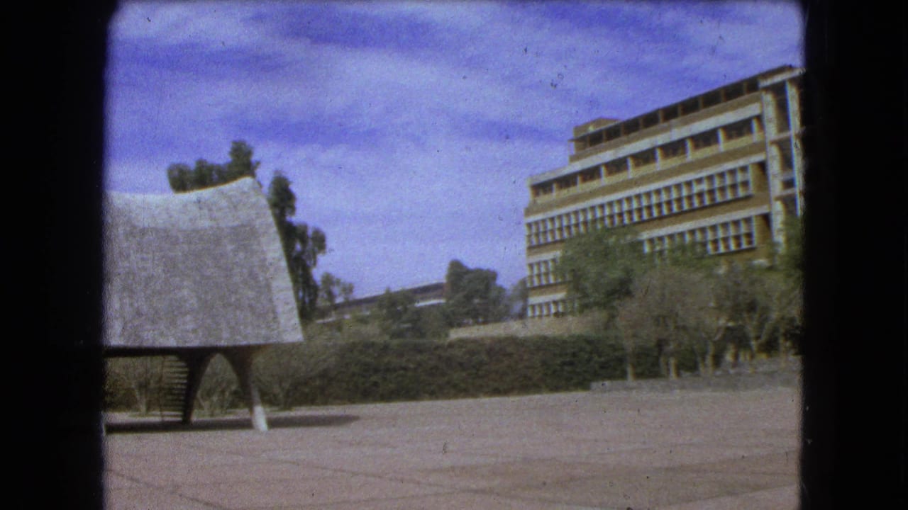 1962 Mexico City frame with a sculptural curved-roof structure in the foreground and a modernist building beyond trees and hedges.