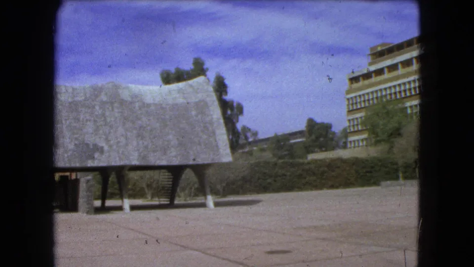 1962 Mexico City home-movie frame showing the textured curved structure close to camera, plaza paving, trees, and part of a modernist facade.