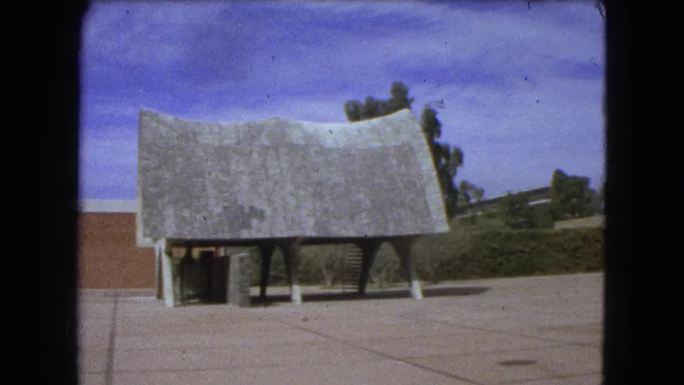 1962 Mexico City 8mm frame centering the sculptural curved-roof structure on a broad paved plaza with trees, hedges, and a building edge.