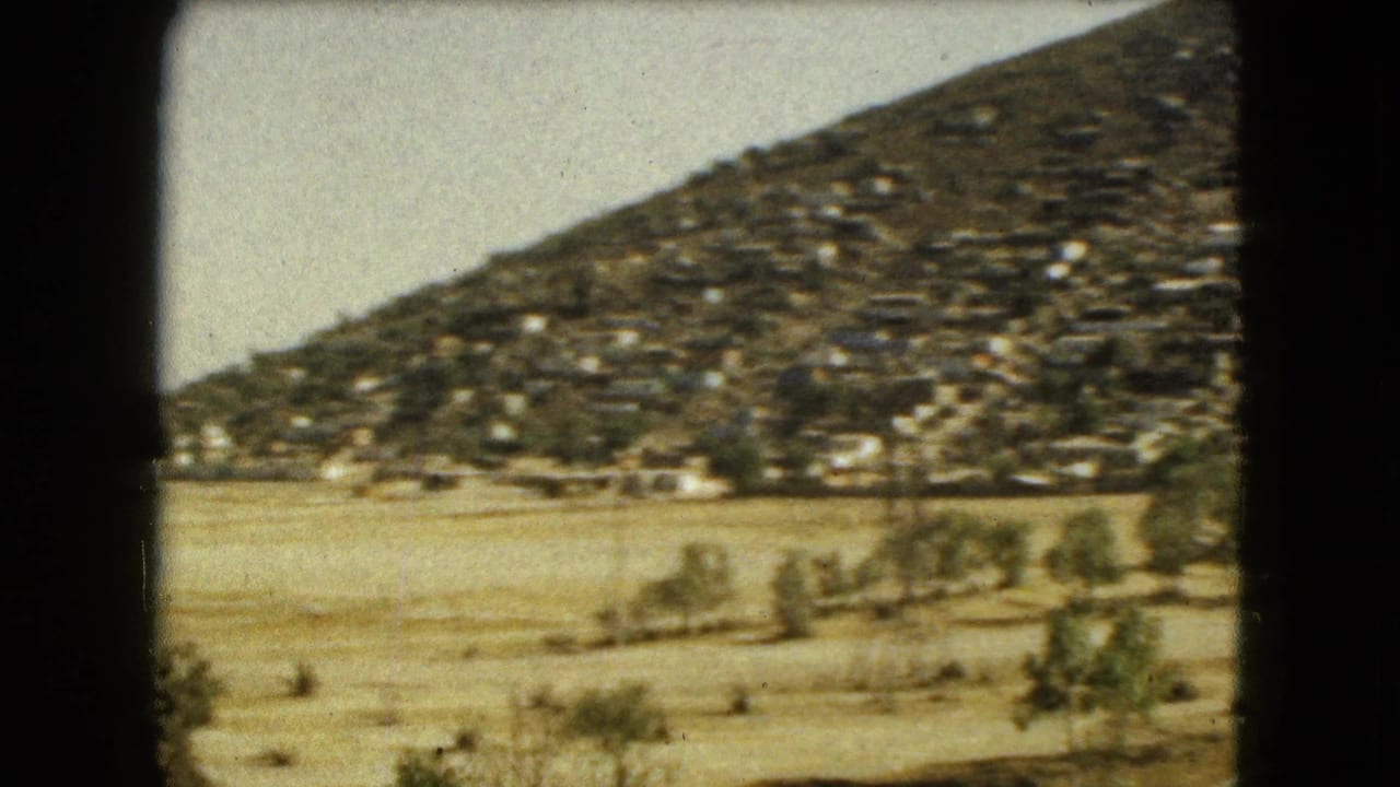 1962 Mexico City 8mm frame showing a hillside covered with small homes beyond dry open ground and sparse trees.