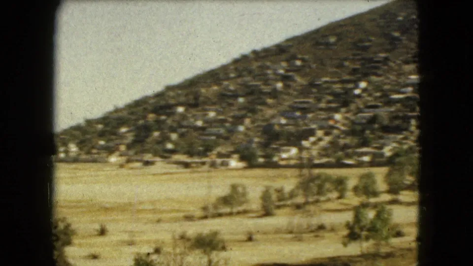 1962 Mexico City 8mm frame showing a hillside covered with small homes beyond dry open ground and sparse trees.