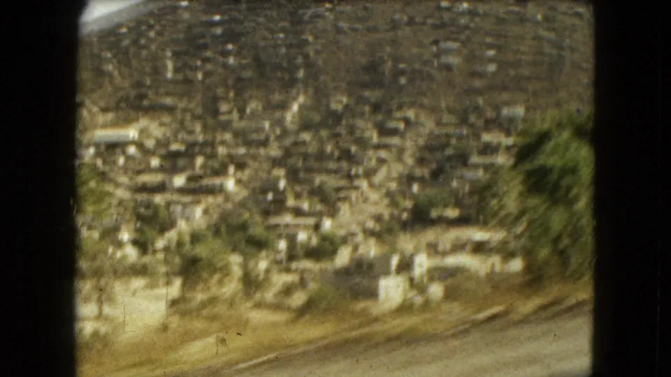 1962 Mexico City frame with a soft moving view of hillside homes, roadside ground, trees, and dry terrain.