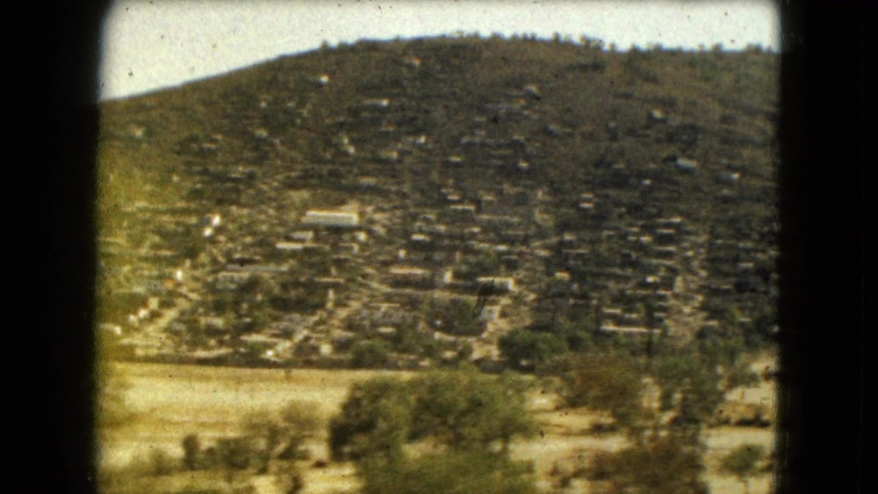 Distant hillside settlement above dry open land with scattered shrubs.