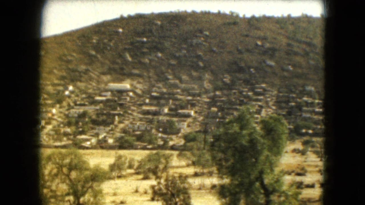 Hillside covered with small structures beyond dry land and trees.