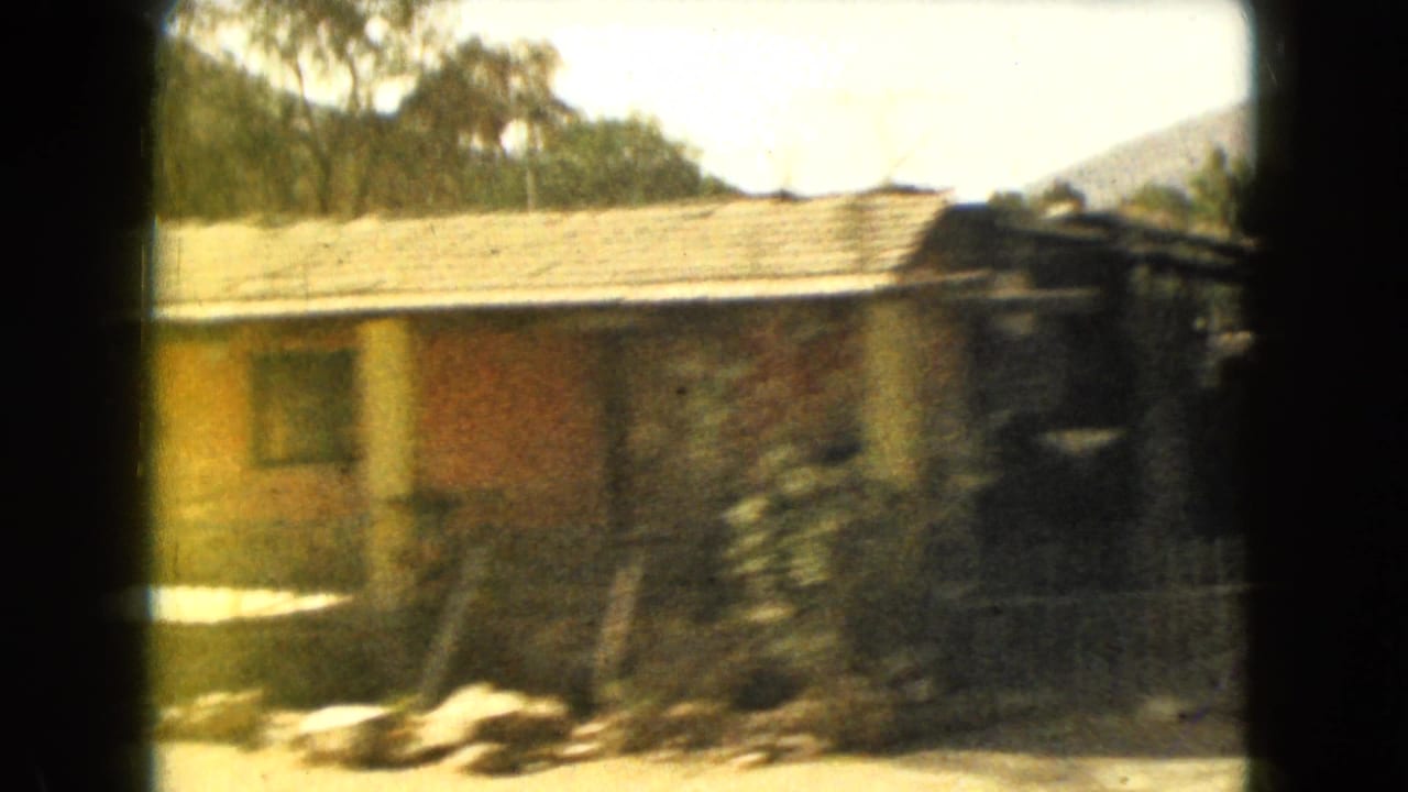 1962 Mexico-area home-movie frame showing a low house with roof tiles, trees, dry roadside ground, and a distant hill.