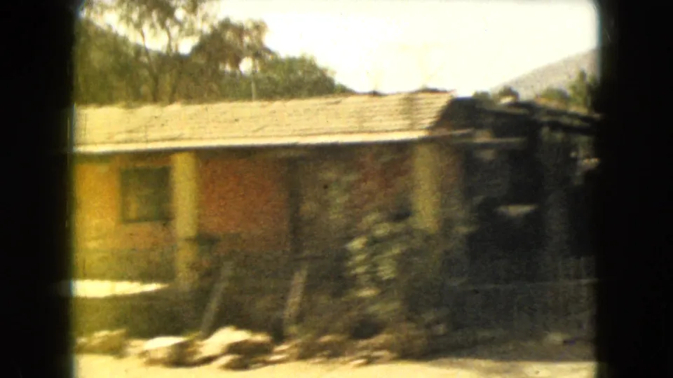 1962 Mexico-area home-movie frame showing a low house with roof tiles, trees, dry roadside ground, and a distant hill.