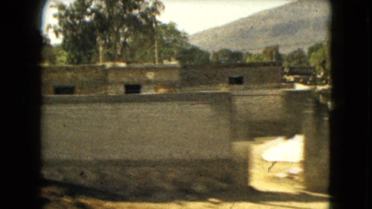 1962 Mexico-area frame with a long wall, simple exterior buildings, trees, dry roadside terrain, and hills in the distance.