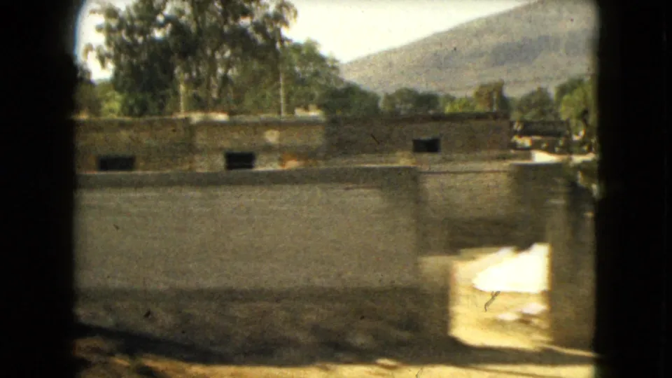 1962 Mexico-area frame with a long wall, simple exterior buildings, trees, dry roadside terrain, and hills in the distance.